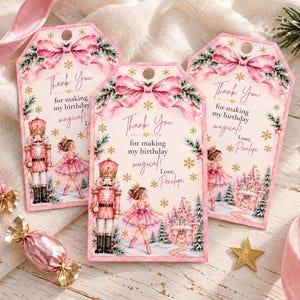 Puede incluir: Tres etiquetas de regalo rectangulares con un lazo rosa, copos de nieve dorados, un cascanueces y una bailarina con tutú rosa. El texto dice: Thank You for making my birthday magical! Love, Penelope. Decoradas con ramas de pino.