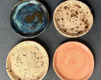 May include: Four handmade ceramic plates in a square arrangement. The plates feature a variety of colors and textures, including a blue and brown glazed plate, two speckled plates, and a coral-colored plate.