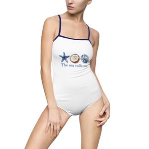 Puede incluir: Traje de baño de una pieza blanco con ribete y tirantes azul marino. Presenta ilustraciones de acuarela de una estrella de mar, un coco y una concha, con el texto "The sea calls me" impreso debajo.