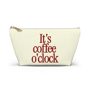 Puede incluir: Un estuche con cremallera de color crema que presenta el texto It's coffee o'clock impreso en una fuente roja oscura. El accesorio tiene un tirador de cremallera de metal dorado en la parte superior y una forma estructurada.