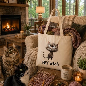 May include: A cream-colored canvas tote bag featuring a graphic of a grumpy, disheveled cat and the text "HEY bitch" printed in black. The bag is filled with various balls of yarn and crochet hooks, with additional yarn and tools on the table.