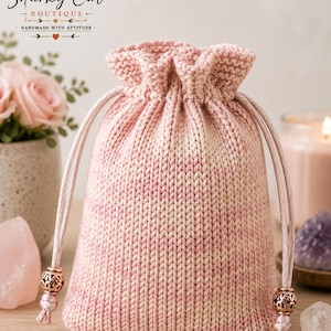 May include: A soft, knitted drawstring pouch in a variegated pale pink and cream yarn. The bag features a gathered top with two matching pink cord ties, each finished with a decorative, intricate metallic rose gold bead at the ends.