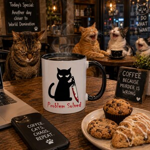 May include: A white ceramic mug with a black handle and rim, featuring a graphic of a black cat holding a bloody knife with the text 'Problem Solved' in red. The mug is placed on a wooden table next to a plate of baked goods and a black smartphone.