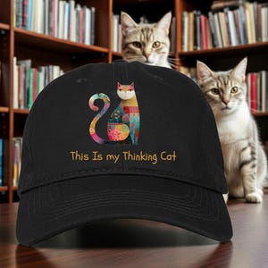 May include: Black baseball cap featuring a colorful cat design with a patterned body and a curled tail. The text "This Is my Thinking Cat" is printed in gold below the cat illustration. The cap is set against a backdrop of bookshelves and cats.