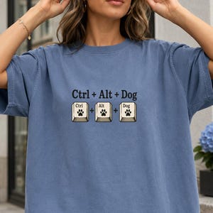 May include: A blue short-sleeve t-shirt featuring a graphic print of three computer keys labeled Ctrl, Alt, and Dog, each displaying a black paw print icon. The text Ctrl + Alt + Dog is printed in black above the three illustrated keyboard keys.