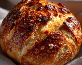 May include: A close-up of a round loaf of bread with a golden-brown crust. The bread has a rustic appearance with a textured surface and is resting on a white cloth. The bread's crust is a mix of light and dark brown.