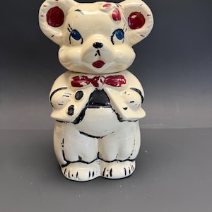 Puede incluir: Una taza de cerámica blanca con asa negra y el texto "But First, Coffee" en negrita negra. La taza se encuentra sobre un fondo borroso, sugiriendo un ambiente acogedor.