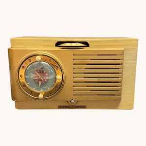 Peut inclure: Radio General Electric vintage de couleur or avec un cadran d'horloge. La radio comporte une horloge ronde avec un fond bleu clair et des chiffres et aiguilles dorés. La radio a une grille de haut-parleur et un bouton de réglage. Les mots "GENERAL ELECTRIC" sont imprimés en bas.