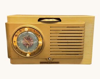 May include: Vintage gold-colored General Electric radio with a clock face. The radio features a round clock with a light blue background and gold-colored hands and numbers. The radio has a speaker grill and a tuning dial. The words "GENERAL ELECTRIC" are printed on the bottom.