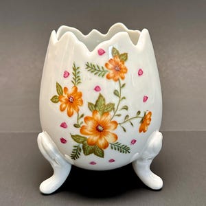 May include: White ceramic egg-shaped container with a scalloped rim and three small feet. The container is decorated with orange flowers, green leaves, and pink accents. A decorative piece for the home.