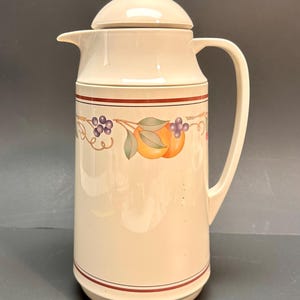 Könnte beinhalten: Cremefarbener Keramikkrug mit Deckel und Henkel. Der Krug ist mit einem horizontalen Band mit Abbildungen von Trauben und Orangen verziert. Ein rot-brauner Streifen akzentuiert die Ober- und Unterseite.