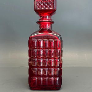 May include: A ruby red glass decanter with a square body and a matching stopper. The decanter features a grid-like pattern on the body and stopper. The stopper has a geometric design. The decanter is a vintage style.