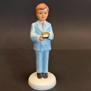 Könnte beinhalten: Keramikfigur eines Jungen in hellblauem Anzug, der ein kleines Buch hält. Die Figur hat rotes Haar und steht auf einem weißen Sockel mit dem Text "My First Communion".