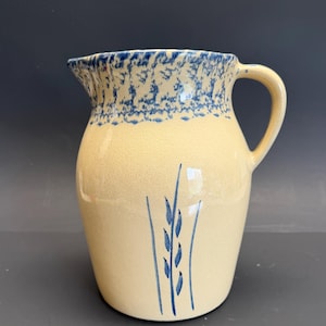 Op de afbeelding: Een vintage keramische kan met een crèmekleurig lichaam en een blauw gespikkelde rand. De kan heeft een blauw tarwe-aren-ontwerp op de voorkant en een gebogen handvat. Deze decoratieve kan is geschikt voor het serveren van dranken.