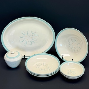 May include: A ceramic dinnerware set featuring an oval platter, a large bowl, a lidded sugar bowl, two medium bowls, and two small bowls. Each piece is cream-colored with a light blue rim and a central pattern of blue swirls and gold stars.