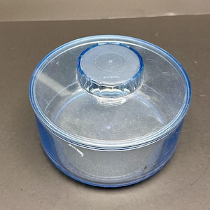 Pode incluir: Um recipiente redondo e transparente com uma borda azul e uma tampa azul a condizer. A tampa tem um botão decorativo facetado. O recipiente é feito de um material transparente.