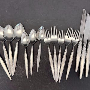May include: A set of stainless steel flatware including six spoons of varying sizes, five forks, and four knives. Each piece features a slender handle with a textured, decorative border pattern along the edges and a polished, reflective finish.