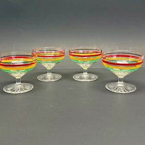 Könnte beinhalten: Vier klare Champagner-Coupe-Gläser mit farbigen horizontalen Streifen in Rot, Gelb, Grün und Schwarz. Die Gläser haben eine runde Schale und einen kurzen Stiel mit dekorativem Fuß. Der Hintergrund ist einfarbig dunkelgrau.