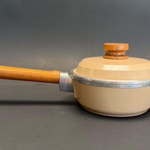 May include: Tan-colored vintage saucepan with a wooden handle and lid. The handle is attached with a metal bracket. The lid has a wooden knob. The pan has a silver rim. A small metal loop is at the end of the handle.