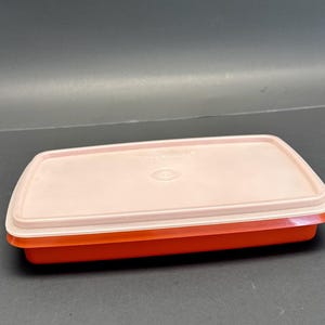 May include: A rectangular, vintage Tupperware container with a coral-colored base and a light pink lid. The lid has a subtle, rounded design and a small circular indentation in the center. The container is made of durable plastic.