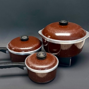 Peut inclure: Un ensemble de trois ustensiles de cuisine émaillés marron avec des bords argentés et des poignées noires. La collection comprend deux casseroles à long manche et une grande marmite, toutes avec des couvercles marron assortis.