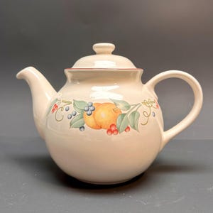 May include: Cream-colored ceramic teapot with a lid and handle. The teapot is decorated with a colorful fruit and leaf design, featuring oranges, blueberries, and red berries. The rim of the lid has a thin red line.