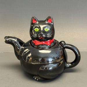 May include: Black ceramic teapot shaped like a cat. The cat has green eyes, red ear interiors, and a red bow tie. The teapot has a handle and a spout. The teapot is on a dark surface.
