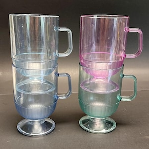 Puede incluir: Dos pilas de tazas de plástico transparente en tonos azul, rosa y verde. Cada taza tiene un asa y una base de pedestal. Las tazas son transparentes, permitiendo el paso de la luz, y están dispuestas sobre un fondo oscuro.