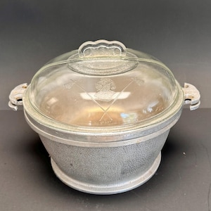 May include: A vintage, round, lidded casserole dish with a clear glass top and a textured, silver-colored metal base. The glass lid features an etched design of crossed golf clubs and a crest. The dish has side handles.