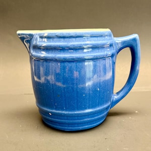Könnte beinhalten: Ein blauer Keramikkrug mit einem fassartigen Design, das vertikale Rillen und horizontale Bänder aufweist. Der Krug hat einen ausgeprägten Ausguss auf der linken Seite und einen stabilen, kantigen Griff auf der rechten Seite mit Glasur.