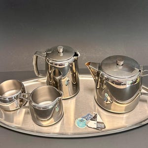 Può includere: Un servizio da tè in acciaio inossidabile su un vassoio ovale. Il set comprende una teiera, una caffettiera, una lattiera e una zuccheriera. Il set ha una superficie lucida e riflettente. Un'etichetta è visibile sul vassoio.