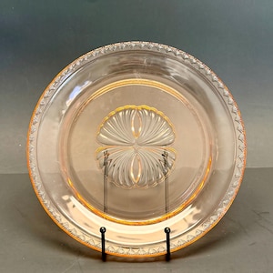 May include: A clear, pink glass plate with a decorative rim and a central design. The plate has a gold-colored trim and a stylized floral pattern in the center. The plate is displayed on a black metal stand.