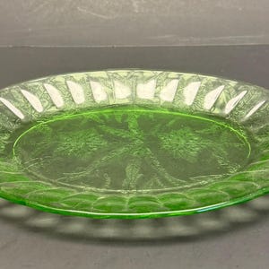 Puede incluir: Un plato de vidrio verde claro transl&uacute;cido con un patr&oacute;n floral prensado en el centro y un borde festoneado con nervaduras verticales. El plato circular tiene una superficie lisa y reflectante con un tono verde vibrante y distintivo.