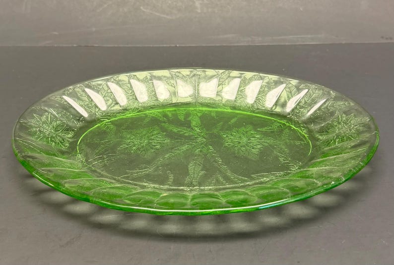 Puede incluir: Un plato de vidrio verde claro transl&uacute;cido con un patr&oacute;n floral prensado en el centro y un borde festoneado con nervaduras verticales. El plato circular tiene una superficie lisa y reflectante con un tono verde vibrante y distintivo.