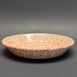Könnte beinhalten: Eine runde, flache Schale mit einem gesprenkelten Muster in Rot und Creme. Die Keramikschale hat einen leicht erhöhten Rand und eine glatte Innenfläche. Das gesprenkelte Design bedeckt die gesamte Oberfläche der Schale.