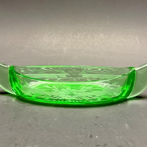 May include: A bright green glass dish in a boat shape. It has etched floral patterns on the base and sides, with curved handles at each end. The glass is translucent, giving it a delicate appearance. The dish is likely for decorative use.
