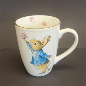 May include: White ceramic mug featuring a watercolor illustration of a rabbit in a blue jacket reaching for a pink butterfly. The rim is trimmed with gold. Two additional pink butterflies are near the rim.