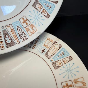 Könnte beinhalten: Zwei weiße Keramikplatten mit einem braunen und blauen geometrischen Muster am Rand. Das Design umfasst Formen wie Blätter, Kreuze und Sternausbrüche. Die Teller haben eine moderne Ästhetik aus der Mitte des Jahrhunderts.