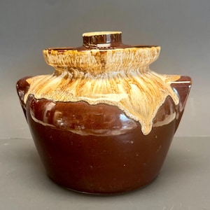 Può includere: Un vaso di fagioli in ceramica marrone e beige vintage con coperchio. Il vaso ha un corpo arrotondato con due manici e un coperchio con un piccolo pomello. La glassa ha un effetto gocciolante, con colori beige e marrone.