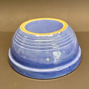 Puede incluir: Un cuenco de cerámica azul con un borde amarillo claro. El cuenco tiene forma redondeada con estrías horizontales alrededor del exterior. El cuenco es de color azul liso con una textura moteada.