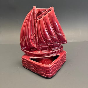 May include: A maroon ceramic sailboat-shaped planter. The planter features a detailed sailboat design with a large sail and a rectangular base with a wavy pattern. The interior of the planter is visible.