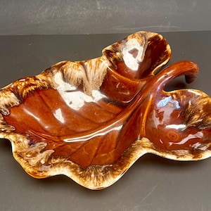 Peut inclure: Un plat en céramique marron en forme de feuille avec une finition brillante. Le plat a une poignée et une palette de couleurs bicolores, avec une bordure marron clair et blanche sur les bords. Le plat est probablement vintage.