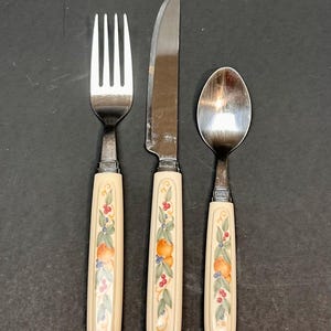 May include: A three-piece silverware set featuring a fork, knife, and spoon. The handles are off-white with a painted design of oranges, berries, and green leaves. The utensils are arranged on a dark surface.