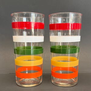 Op de afbeelding: Twee heldere drinkglazen met horizontale banden in rood, wit, groen, geel en oranje. De glazen zijn cilindrisch en lijken van glas te zijn. De banden zijn gelijkmatig verdeeld en creëren een retro-ontwerp.