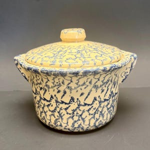 Puede incluir: Una fuente de cerámica para hornear con tapa. La fuente y la tapa son de color crema con un patrón de salpicaduras azul. La fuente tiene dos asas y un pequeño pomo en la tapa. El diseño es rústico y vintage.