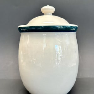 Puede incluir: Un tarro de cerámica blanco con tapa. El tarro tiene forma redondeada y una banda verde oscuro alrededor del borde. La tapa es blanquecina con un pequeño pomo en la parte superior. Ideal para la cocina.