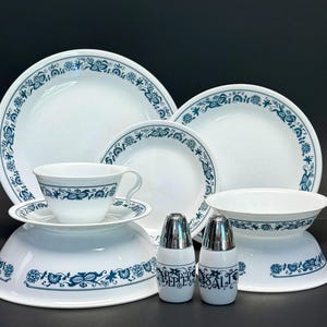 Puede incluir: Un juego de vajilla blanco con un patrón floral azul. La colección incluye platos grandes y pequeños, un cuenco, una taza de té con platillo y dos saleros y pimenteros marcados como "PEPPER" y "SALT" con tapas metálicas plateadas.