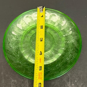 Puede incluir: Un plato redondo de vidrio verde transl&uacute;cido con un patr&oacute;n de hojas en relieve en toda su superficie. Una cinta m&eacute;trica amarilla est&aacute; colocada verticalmente sobre el centro, indicando que el plato tiene un di&aacute;metro de 15,24 cm.