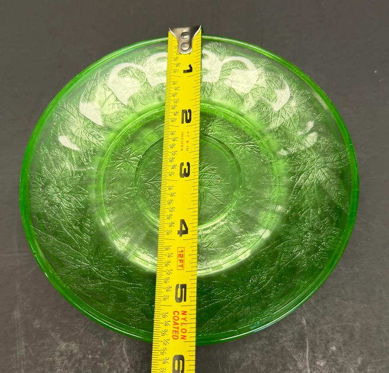 Puede incluir: Un plato redondo de vidrio verde transl&uacute;cido con un patr&oacute;n de hojas en relieve en toda su superficie. Una cinta m&eacute;trica amarilla est&aacute; colocada verticalmente sobre el centro, indicando que el plato tiene un di&aacute;metro de 15,24 cm.