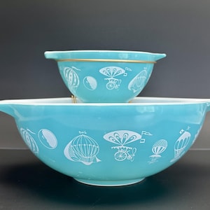 May include: Two turquoise blue mixing bowls with a white design of hot air balloons and a vintage car. The smaller bowl is stacked on top of the larger bowl.